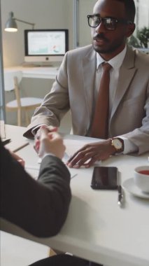 Resmi takım elbiseli genç Afro-Amerikan işadamının dikey çekimi ve ofis masasındaki Kafkas meslektaşıyla el sıkışması.