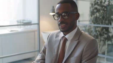 Resmi giyimli ve gözlüklü genç Afro-Amerikalı iş adamının ofiste çalışırken kameraya bakıp gülümsediği bir portre.