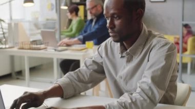 Gündelik kıyafet giyen Afro-Amerikalı bir adam ofiste çalışırken laptop kullanıyor ve belge okuyor.