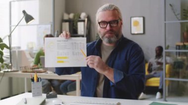 Gündelik kıyafet ve gözlüklü kıdemli işadamı ofiste internette konuşurken kameradaki belgeyi açıklıyor.