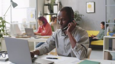 Genç Afro-Amerikalı bir adam, danışmada dizüstü bilgisayar kullanıyor ve destek merkezinde çalışırken internet telefonlarına cevap veriyor.