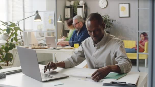 Afroamerykański Biznesmen Luźnym Stroju Piszący Laptopie Czytający Dokumenty Podczas Pracy — Wideo stockowe