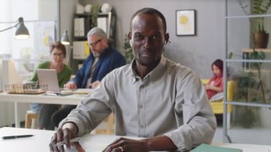 Kablosuz kulaklıklı Afro-Amerikalı adam kameraya el sallıyor, belgeyi gösteriyor ve ofiste çalışırken online video görüşmesinde detayları anlatıyor.