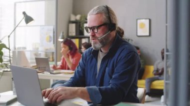 Kablosuz kulaklık takan, dizüstü bilgisayarda yazan ve çağrı merkezinde çalışırken internet üzerinden konuşan kıdemli bir adam.