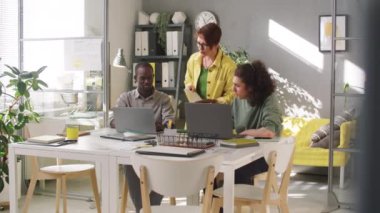 Orta yaşlı kadın yönetici ofiste uyanıyor ve laptoplarda Afro-Amerikalı ve beyaz erkek stajyerlere bir şeyler anlatıyor.