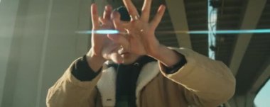 Güneşli bir günde şehir köprüsünün altında dans ederken kameraya bakıp el hareketleri yapan genç hip hop dansçısının anamorfik görüntüsünü yakınlaştır.