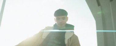 Düşük açılı anamorfik çekim. Beyaz bir adam kameraya bakıyor ve şehrin köprüsünün altında hip hop dansı yapıyor.