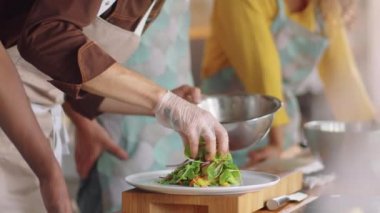 Önlüklü ve eldivenli bir erkek aşçının tabakta salata servis ederken yemek pişirme ustası sınıfına verdiği pozu kaldır.