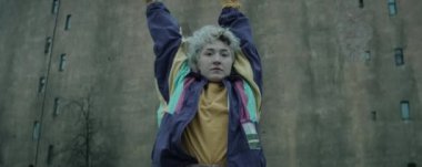 Eski model ceketli genç bir kadının eski beton binanın önünde kameraya bakıp dans ederken anamorfik görüntüsünü yakınlaştır.