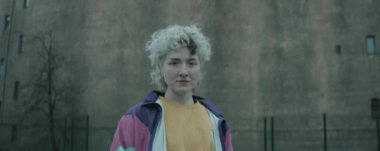 Eski model ceketli genç kadının eski beton binanın önünde dikilip kameraya bakarken anamorfik görüntüsünü yakınlaştır.