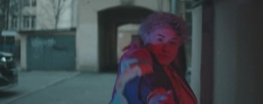 Klasik bombacı ceketi giymiş genç bir kızın kameraya doğru yürüdüğü ve avluda kırmızı ışıkla dans ettiği anamorfik bir görüntü.