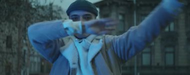 Genç profesyonel erkek dansçının kendine güvenen bir şekilde kameraya baktığı ve akşam dışarıda dururken kol hareketleri yaptığı anamorfik bir çekim.