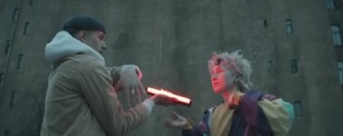 Modayı takip eden sokak kıyafetleri içindeki genç kadın ve erkeğin açık havada dans ederken kırmızı lambanın önünden geçişlerinin anamorfik görüntüsü.