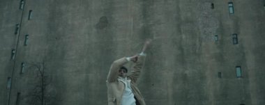 Modayı takip eden ceketli ve şapkalı bir gencin kameraya bakıp eski beton binanın önünde vogue dansı yaptığı belden aşağısı anamorfik bir çekim.