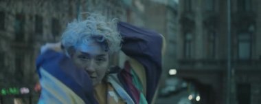 Klasik bombardıman ceketli genç bayan dansçının akşam saatlerinde kameraya bakıp açık havada dans ederken, yan tarafında yanıp sönen renk ışıklarıyla anamorfik görüntüsü.