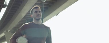 Futbol topu tutan ve şehir köprüsünün altında yürüyen yakışıklı genç sporcunun anamorfik pozunu çek.