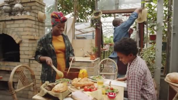 アフリカ系アメリカ人の父親と息子はテーブルに歩いて行き 母親は皿の上に食べ物を置き 温室で家族の夕食で彼らとおしゃべりをする — ストック動画