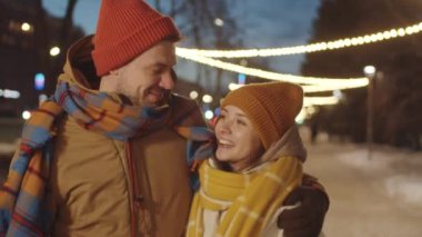 Sokakta Noel ışıklarıyla süslenmiş romantik gece yürüyüşleri yaparken kucaklaşan, gülümseyen ve sohbet eden genç çiftler.