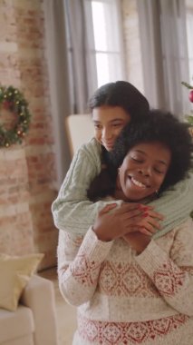 Mutlu Afro-Amerikan kızının dikey portresi neşeli annesine sarılıyor ve evde Noel ağacının yanında birlikte poz verirken kameraya gülümsüyor.