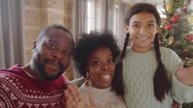 Neşeli Afro-Amerikan ailesi evde Noel ağacının yanında dururken çevrimiçi video aracılığıyla el sallıyor, gülümsüyor ve konuşuyor.