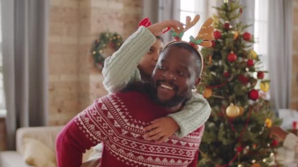 幸せなアフリカ系アメリカ人の女の子座っているお父さんの後ろに彼の鹿アントラーヘッドバンドと遊んでいる間お母さん飾るクリスマスツリーに家 — ストック動画