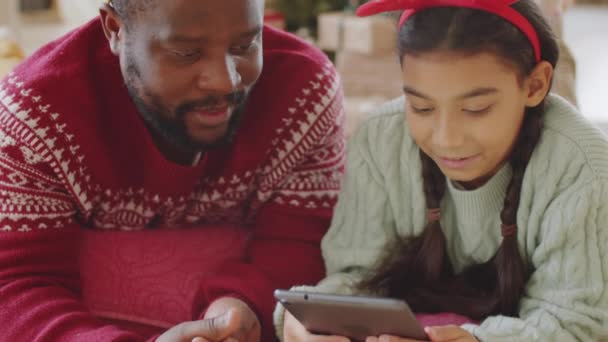 自宅の床の上に一緒に横たわって 笑顔とデジタルタブレット上の何かについてチャットクリスマスヘッドバンドでアフリカ系アメリカ人の父と娘のショットを傾け — ストック動画