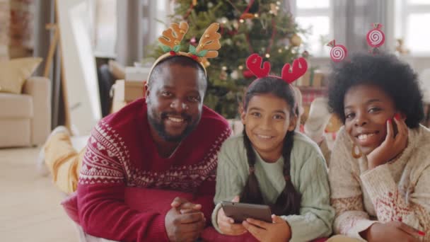 クリスマスのヘッドバンドの幸せなアフリカ系アメリカ人の家族の肖像は デジタルタブレットで床に横になり カメラを見て笑っています — ストック動画