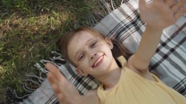 Küçük sevimli bir kızın yaz gününü dışarıda battaniyeye uzanmış kameraya gülümseyen ellerini uzatarak geçirdiği tepeden tırnağa bir portre resmi.