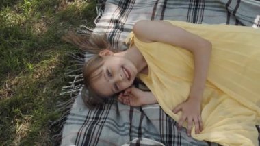 Sarı elbiseli küçük kızın yüksek açılı görüntüsü. Parkta battaniyede dinleniyor. Kameraya gülümsüyor.