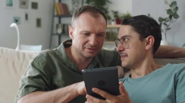 Orta ölçekli bir yakın çekim. Yetişkin, aşık erkeklerin oturma odasındaki kanepede oturup dijital tablette internetten bir şeyler izlediği görüntüler.