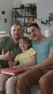 Mutlu, evli, eşcinsel bir çiftin oturma odasındaki kanepede oturup kameraya baktığı orta boy bir fotoğraf.