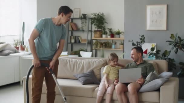 英俊的白人男人在客厅里做家务活 而他的丈夫和女儿则坐在沙发上看笔记本电脑上的东西 — 图库视频影像