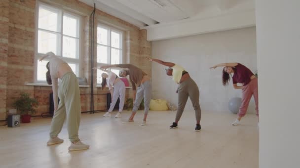 若い多民族の男性と女性のスポーツウェアのグループは スタジオでダンスクラスの準備をしながら 女性コーチとサイドストレッチ運動を行う — ストック動画