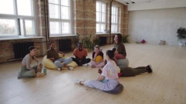 Stüdyoda yerde dinlenen ve dans dersinden sonra sohbet eden genç kadın ve erkeklerin geniş açılı görüntüleri.