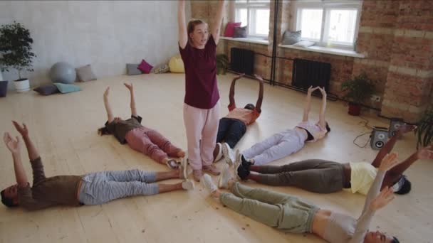 若い多民族の人々が腕を上げて手を動かしながら床の上に横になり 女性教師がその間に立ち スタジオでダンス教室中に動きを見せる — ストック動画