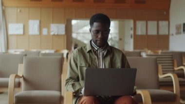 Gözlüklü ve günlük giysili Afro-Amerikalı adam kütüphane salonunda oturuyor ve dizüstü bilgisayarla çalışıyor.