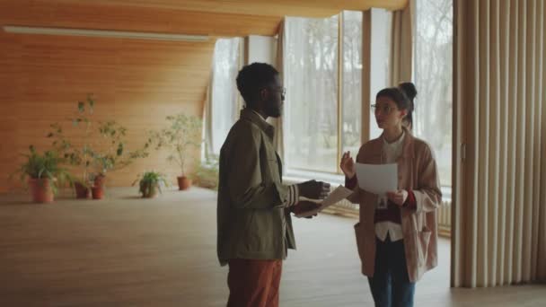 年轻的白人女人和黑人男人拿着文件 一边讨论着什么 一边站在有波浪形天花板的现代礼堂里 — 图库视频影像