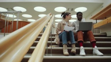 Çok ırklı genç kadın ve erkeğin konuştuğu, elinde kağıt olan ve dizüstü bilgisayarla kütüphanede oturup proje üzerinde çalışırken yakınlaştırın.