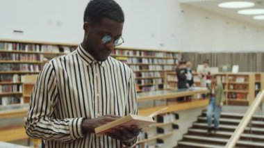 Seçici odak noktası, gözlüklü Afro-Amerikan bir adamın kütüphanede durup kitap okuması.