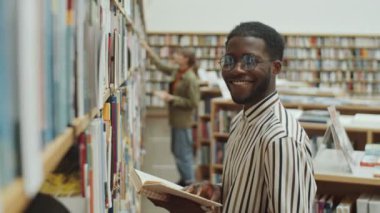 Gözlüklü, çizgili gömlekli, kitap tutan ve kütüphanede raflarda poz verirken kameraya gülümseyen neşeli Afro-Amerikan adam portresi.