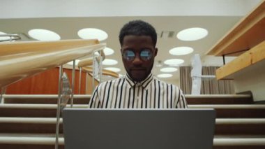 Kütüphanede merdivenlerde oturmuş dizüstü bilgisayarla çalışan gözlüklü Afro-Amerikan bir adamın arkası.