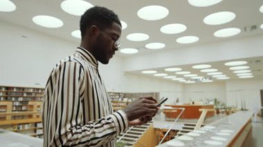 Kütüphanede dikilen ve akıllı telefondaki mobil interneti karıştıran Afro-Amerikalı adamın belden yukarı çekimi.