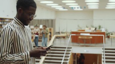 Sırıtan Afro-Amerikan bir adamın kütüphanede durup cep telefonuyla mesajlaşmasını belden yukarı çek.