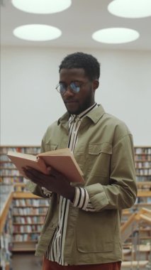 Gözlüklü ve çizgili tişörtlü Afro-Amerikan adamın kütüphanede durup kitap okuduğu dikey bir fotoğraf.