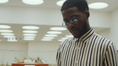 Gözlüklü ve çizgili tişörtlü Afro-Amerikalı adamın kütüphanede durup kitap sayfalarını çevirirkenki görüntüsü.