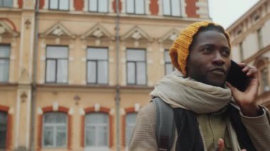 Düşük açılı çekimde, Afrika kökenli Amerikalı erkek turist dış giyim ve örgü şapka ile eski kasabada dikilip telefonda sohbet ederken görülüyor.