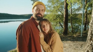 Genç romantik çift göl kıyısında durup kameraya poz verirken yaz tatilini doğada geçiriyorlar.