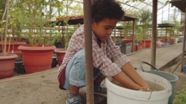 Küçük Afro-Amerikalı çocuk serada çömelmiş ellerini bir kova suda yıkıyor.