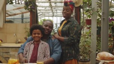 Mutlu Afro-Amerikan aile portresi kameraya bakıyor ve serada yemekte gülümsüyor.