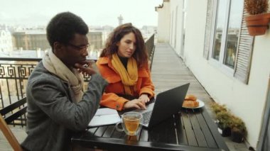 Neşeli Kafkasyalı iş kadını ve Afro-Amerikalı erkek meslektaşı tee ve kruvasanlarla çatıda oturuyor, dizüstü bilgisayar kullanıyor ve konuşuyor.